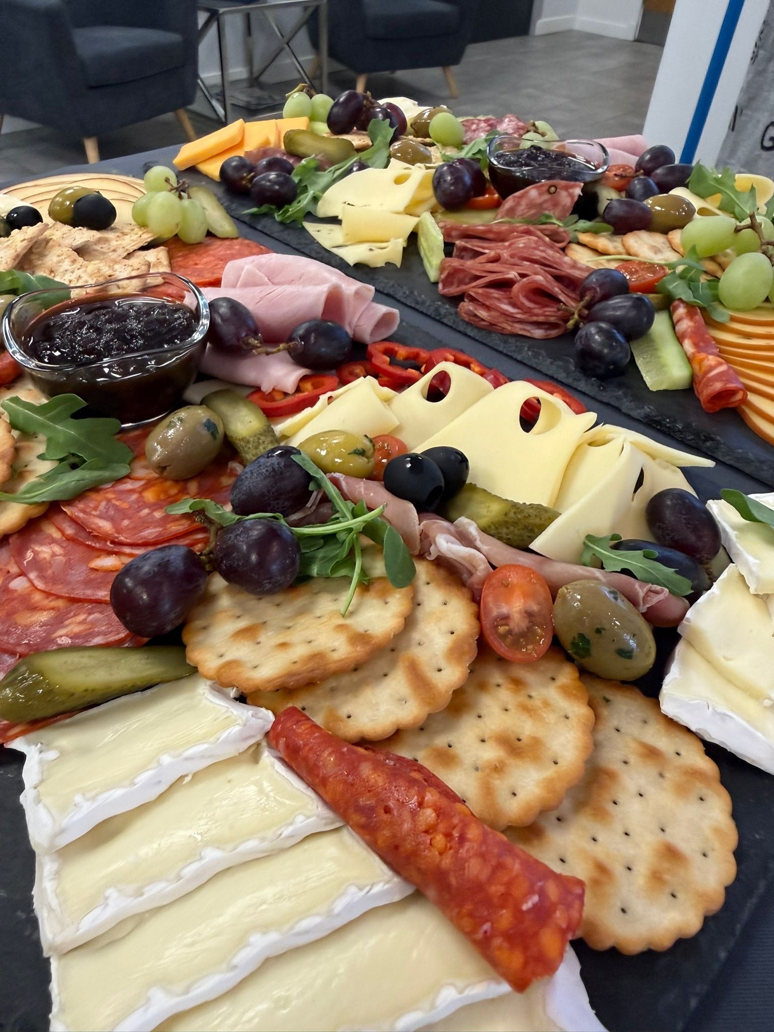 A large charcuterie board spread filled with cheeses, crackers, sliced meats, grapes, olives, pickles, and a small bowl of dark dipping sauce, photographed on a table in an indoor setting.