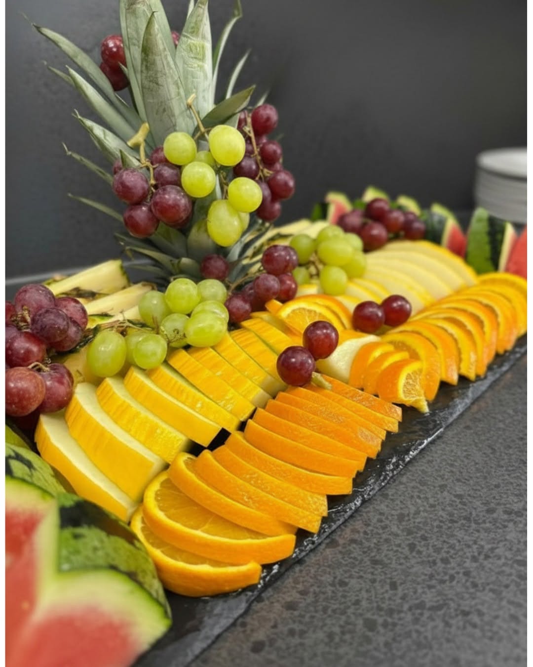 A long black serving platter filled with sliced citrus fruit, green and red grapes, pineapple, watermelon, and other fresh fruit, photographed on a dark countertop against a dark background