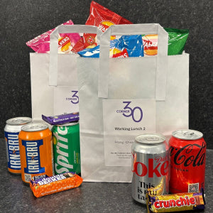 A white paper shopping bag with the 'Corner30' logo, filled with colorful crisp bags, alongside assorted canned fizzy drinks and chocolate bars on a dark textured background.