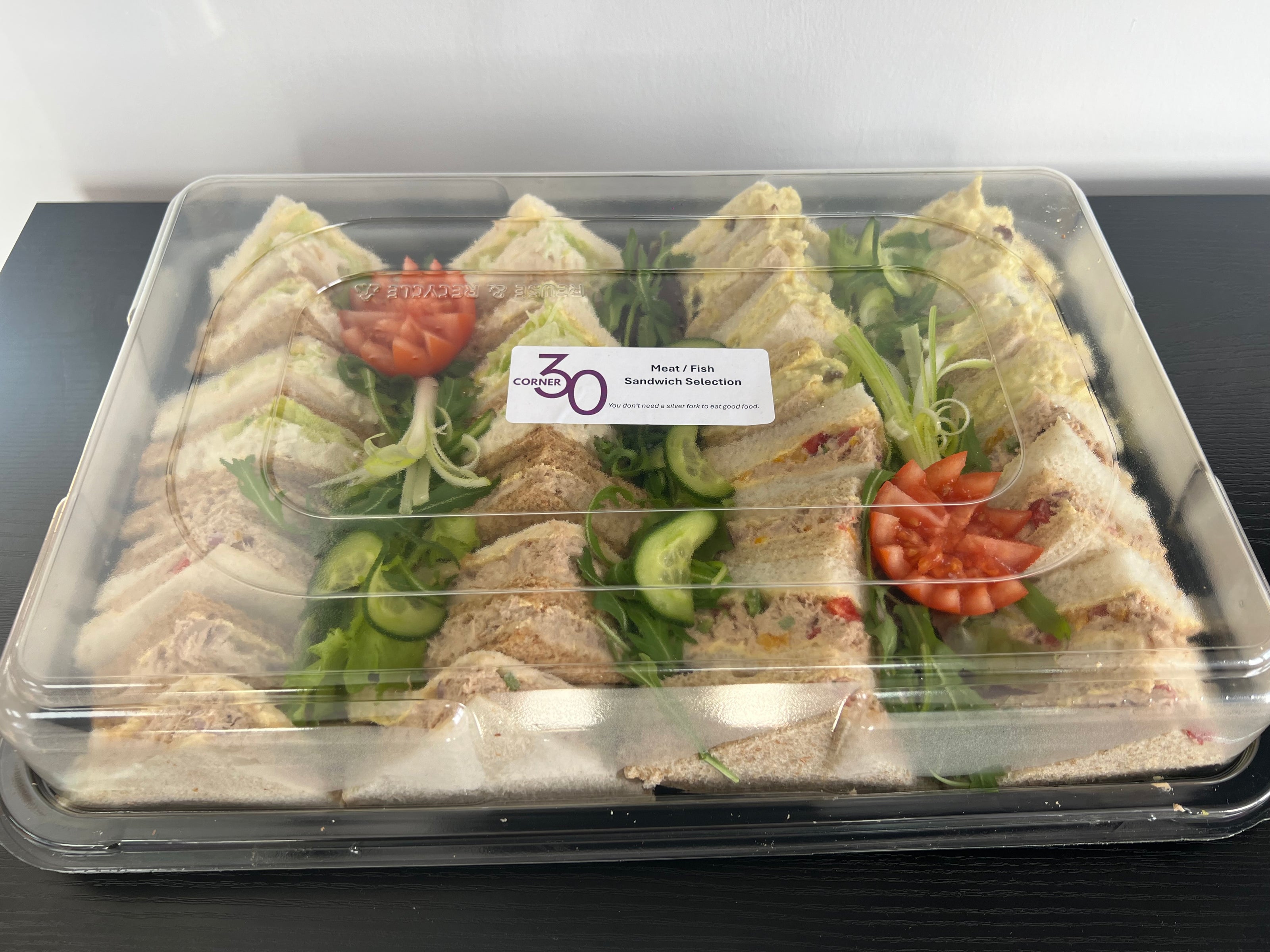 A clear plastic clamshell container holding multiple triangular sandwiches arranged on a bed of green salad, with small tomato garnish pieces. A small white label is visible on the lid, and the food sits on a dark tabletop against a light wall.