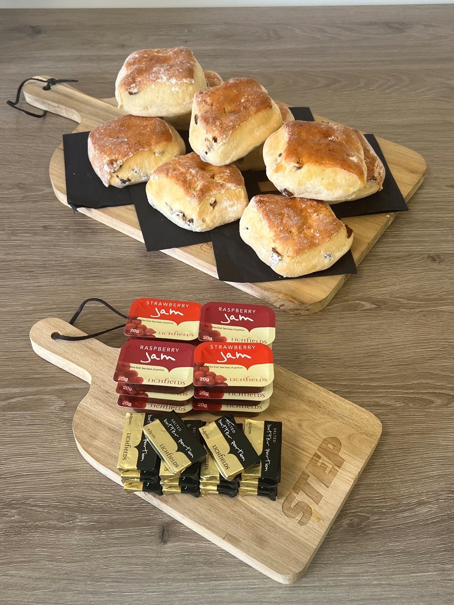 Two wooden serving boards on a wood-grain tabletop, styled with baked scones on one board and packaged butter and jam on the other. The scones are dusted with flour and appear golden brown.
