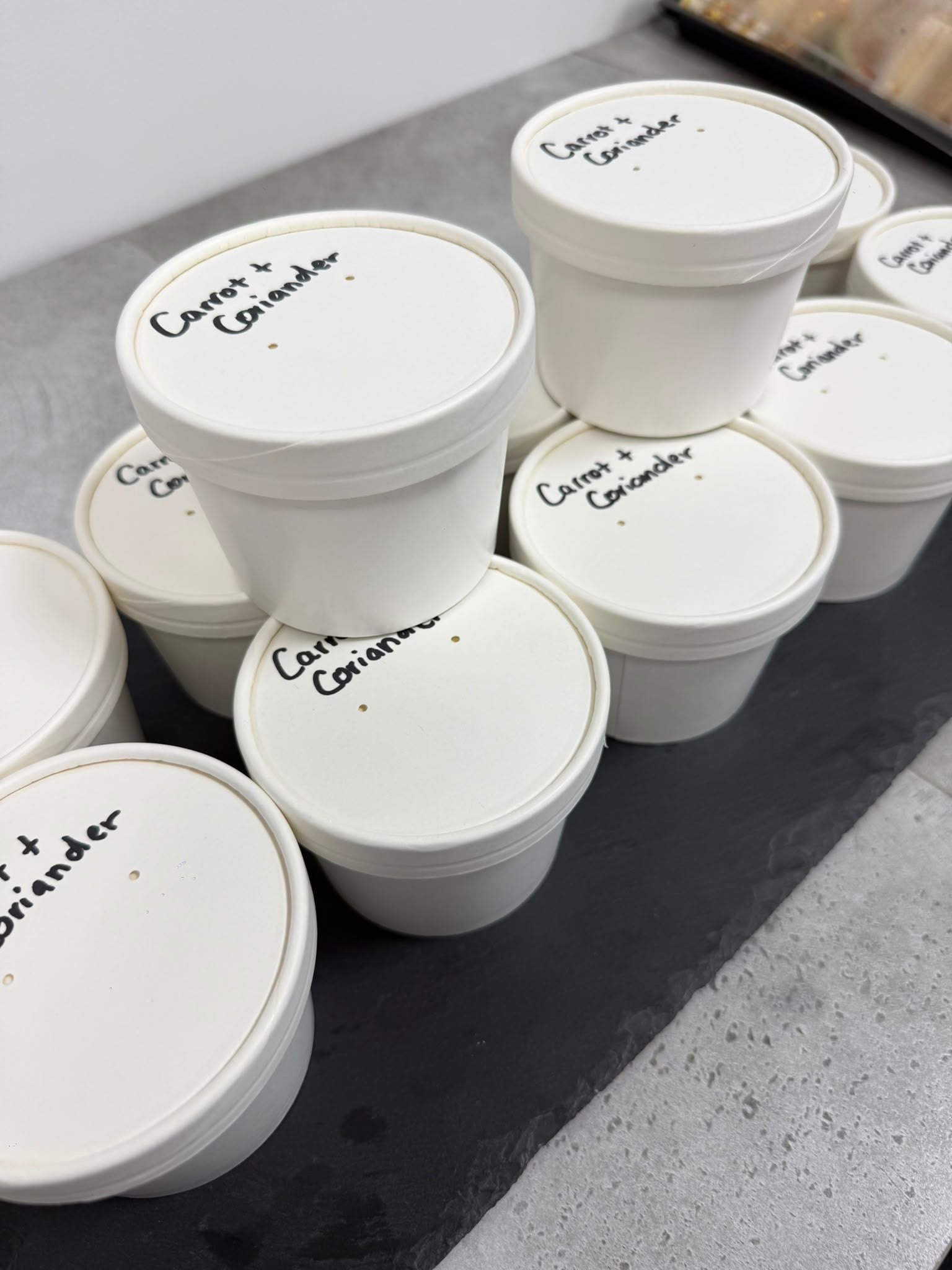A cluster of white plastic food storage containers with lids, each labeled with handwritten text 'Carrot and Coriander'.
