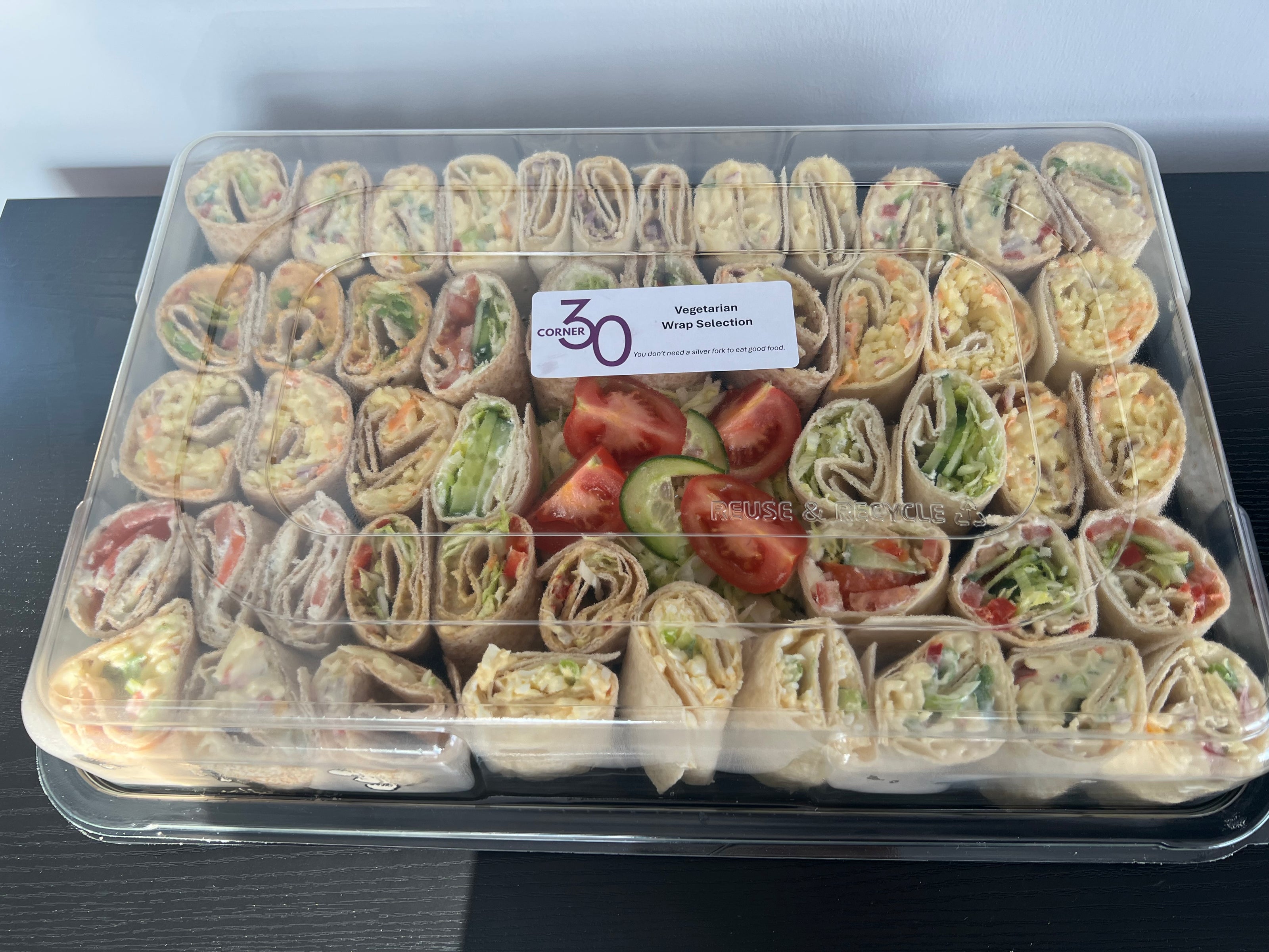 A clear plastic container filled with 30 veggie wrap sandwiches arranged in rows, with sliced tomatoes and cucumber placed in the center. A white label on the lid reads 'Corner30 Vegetarian Wrap Selection'.